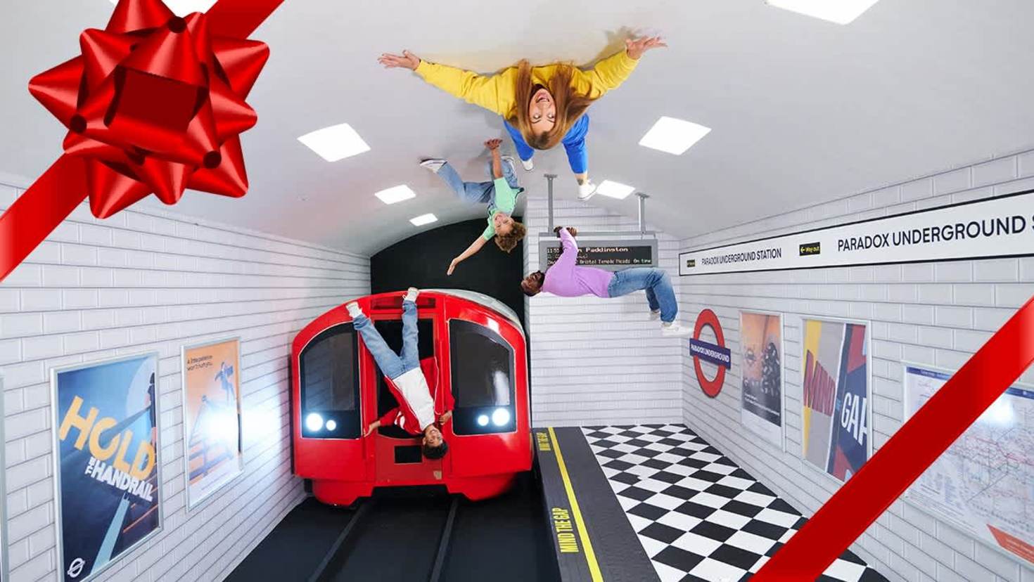 A family appears to hang upside down from the ceiling of a Tube station illusion at Paradox Museum London, with a red London Underground train below and optical illusions on the tiled walls. A family appears to hang upside down from the ceiling of a Tube station illusion at Paradox Museum London, with a red London Underground train below and optical illusions on the tiled walls.