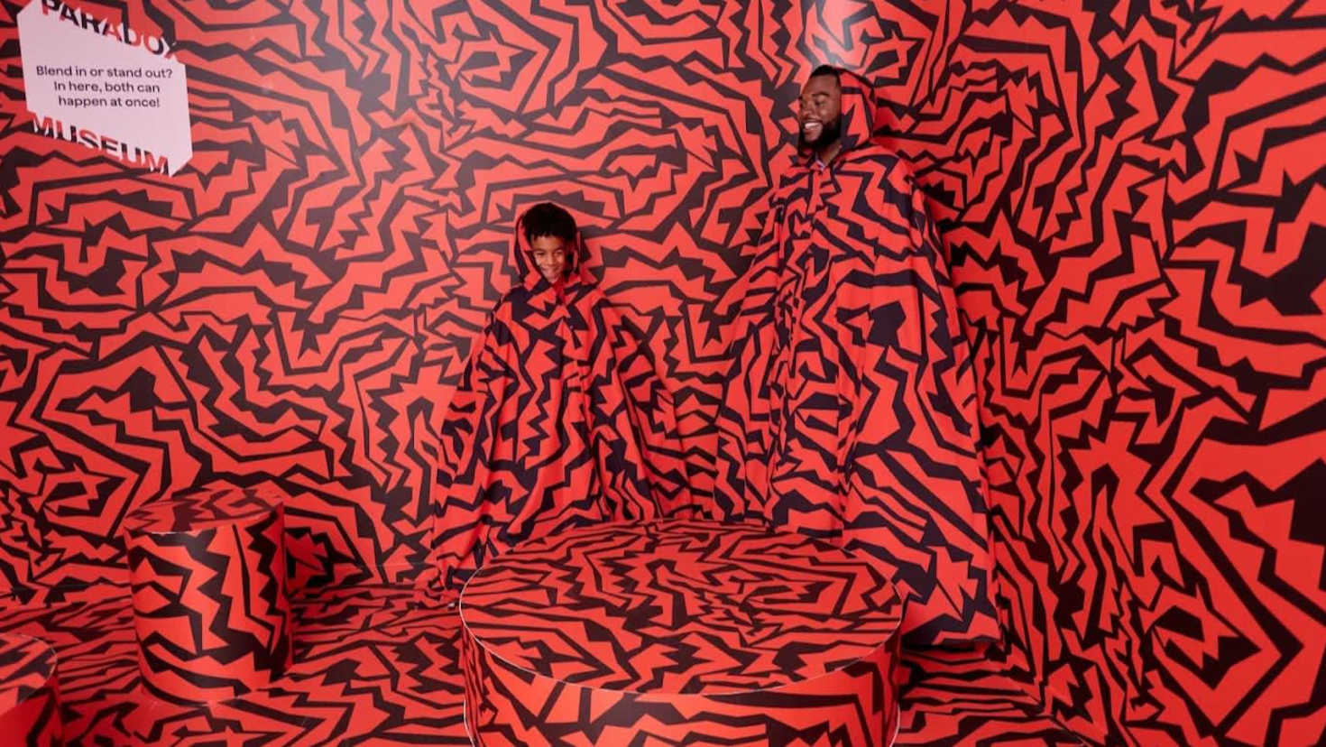 Child and adult blending into patterned red wall at Paradox Museum, creating an optical illusion with matching cloaks.
