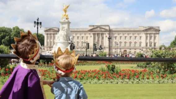 Two children wearing crowns looking at Buckingham Palace from the gardens.