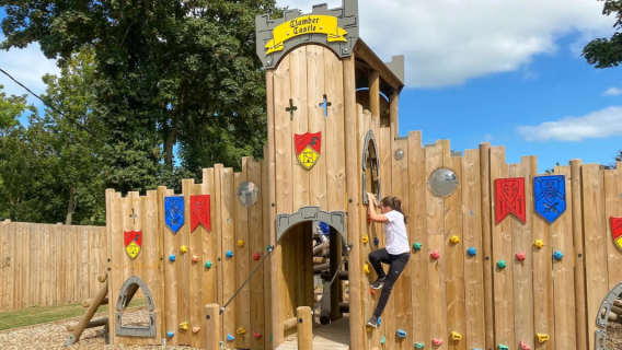 A child climbs a wooden castle-themed playground structure decorated with colorful shields.