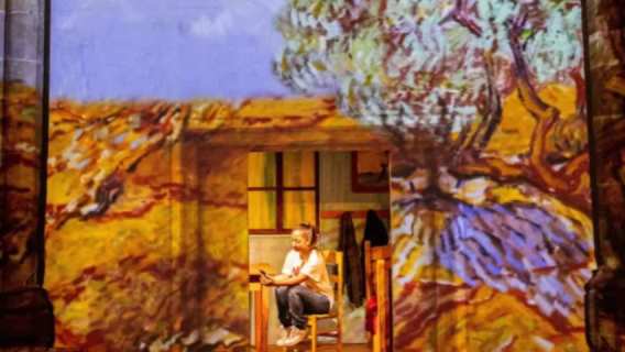 A woman sits in a chair reading inside a room that blends into a large-scale Van Gogh landscape projection, with vivid yellow fields and swirling trees enveloping the scene.