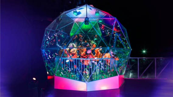 A group of players inside the iconic Crystal Dome scramble to catch swirling golden tokens under bright neon lights. The glass dome glows with vibrant greens, purples, and pinks.