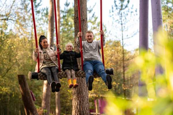 A family of three are on a large wooden swing in the middle of a forest adventure park. Surrounded by tall trees and natural woodland, the parents sit on either side of a smiling child in yellow boots, all swinging high with laughter and excitement.