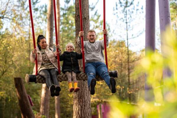 A family of three on a large wooden swing in the middle of a forest adventure park. Surrounded by tall trees and natural woodland, the parents sit on either side of a smiling child in yellow boots, all swinging high with laughter and excitement. A family of three on a large wooden swing in the middle of a forest adventure park. Surrounded by tall trees and natural woodland, the parents sit on either side of a smiling child in yellow boots, all swinging high with laughter and excitement.