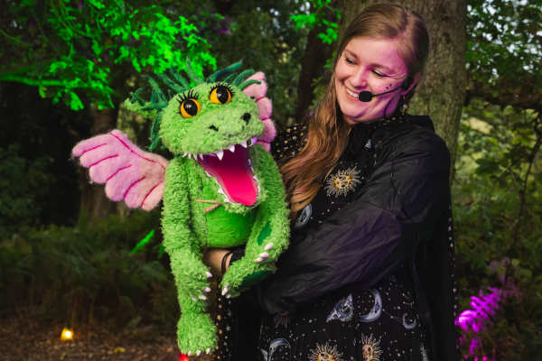 An outdoor performance takes place on a wooden stage at BeWILDerwood. A performer in a pink hoodie and headset microphone gestures animatedly towards a large audience of children and adults, who sit under a wooden canopy among tall pine trees, watching and joining in with the show.
