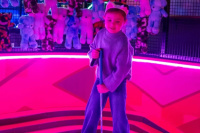 Child holding a golf club in front of a wall of soft toy prizes at a glow-in-the-dark mini golf venue.