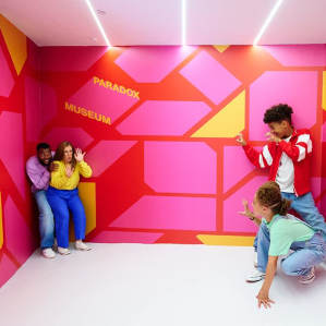 Four children play with perspective in the vibrant Ames Room at Paradox Museum London, where the room's optical illusion makes them appear dramatically different in size and position.