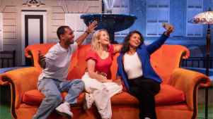 A trio of smiling visitors sits on the iconic orange couch in front of the Friends fountain set, with arms around each other as one holds up a phone for a selfie. The scene recreates the show's famous opening credits moment.