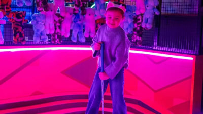 Child holding a golf club in front of a wall of soft toy prizes at a glow-in-the-dark mini golf venue.
