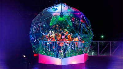 A group of players inside the iconic Crystal Dome scramble to catch swirling golden tokens under bright neon lights. The glass dome glows with vibrant greens, purples, and pinks.