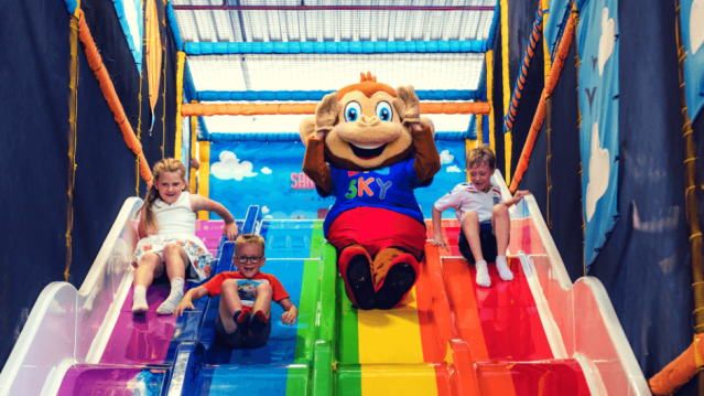 Kids and monkey mascot racing down a four-lane slide at Big Sky Adventure Play