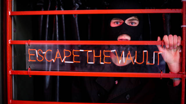 Person in mask looking through sign that says 'Escape The Vault' 