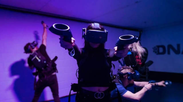 Group of adults wearing VR headsets at DNA VR Manchester, playing multiplayer virtual reality games in a neon-lit room