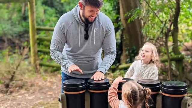 Dad and two daughters enjoy interactive drumming together in a forest play area at Hoo Zoo.