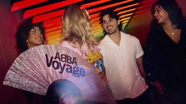 A group of friends, one holding a sparkling “ABBA Voyage” fan, smiles and talks under vibrant tunnel lighting.