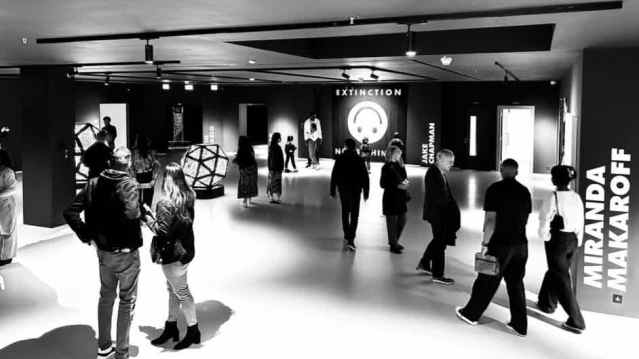 People explore a dark, modern art exhibition with geometric sculptures and illuminated signage reading “EXTINCTION.”