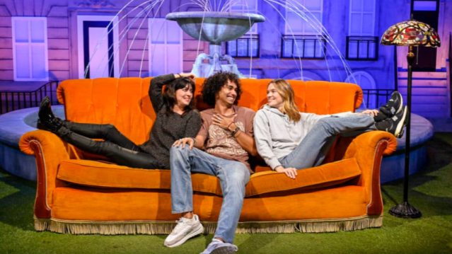 Three friends lounge playfully on the signature orange couch in front of the fountain set, laughing and leaning toward one another. The recreated night-time city backdrop and vintage floor lamp add to the nostalgic feel.