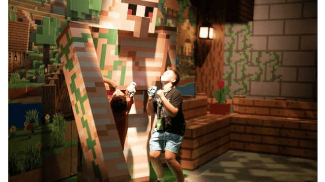 Two kids interact with a giant Minecraft iron golem figure in a themed exhibit area, looking up in awe. The background features classic pixel-style flowers, wooden textures, and a glowing lantern for ambiance.