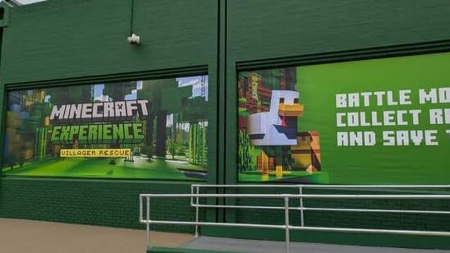 Exterior view of the “Minecraft Experience: Villager Rescue” attraction, featuring promotional signage with a forest background, a chicken, and the tagline “Battle mobs, collect resources, and save the village.”