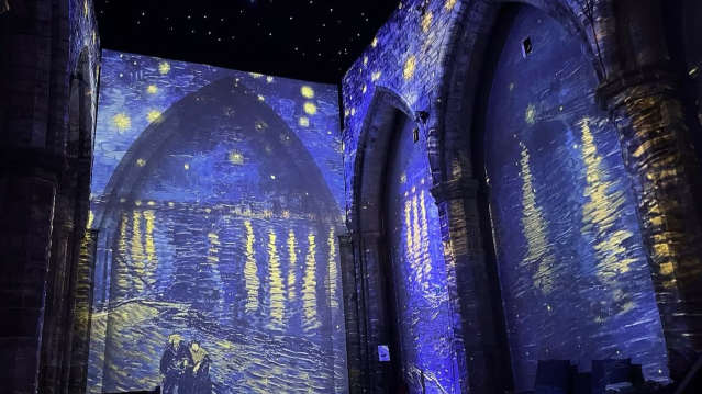 Van Gogh’s Starry Night Over the Rhône projected in deep blue tones with glowing yellow stars reflecting in the water, enveloping the Gothic arches of a cathedral interior.