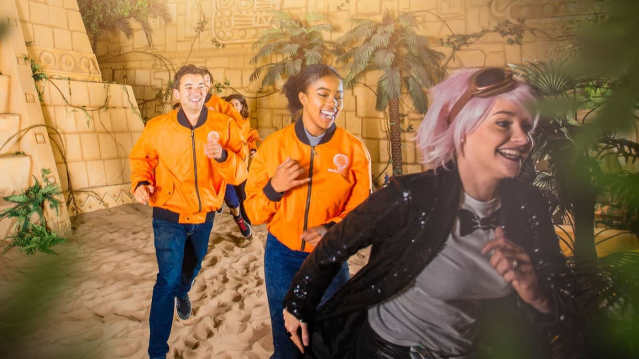 A game host leads a group of players in orange jackets sprinting across a sandy Aztec-themed zone with jungle plants and stone walls.A game host leads a group of players in orange jackets sprinting across a sandy Aztec-themed zone with jungle plants and stone walls.